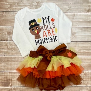 May include: A white long-sleeved baby bodysuit with a turkey graphic and the text "MY ROLLS ARE HOMEMADE." Accompanied by a layered tutu skirt in brown, orange, and yellow, and a large brown bow.