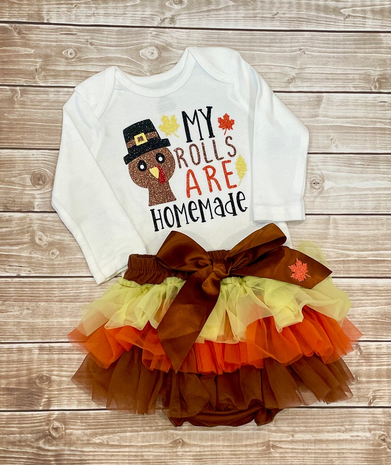 May include: A white long-sleeved baby bodysuit with a turkey graphic and the text "MY ROLLS ARE HOMEMADE." Accompanied by a layered tutu skirt in brown, orange, and yellow, and a large brown bow.