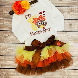 May include: A white long-sleeved onesie with the text "My 1st Thanksgiving" and a glitter turkey design. The onesie is paired with a brown tulle skirt with orange, yellow, and brown layers. A headband with orange and yellow flowers completes the outfit.