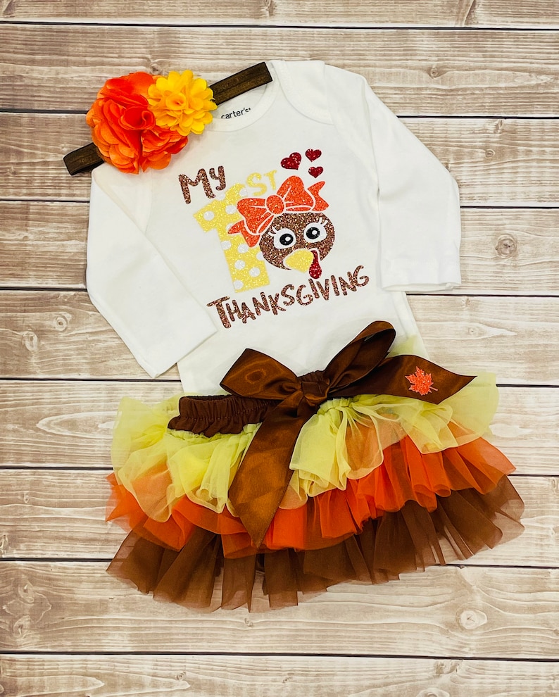 May include: A white long-sleeved onesie with the text "My 1st Thanksgiving" and a glitter turkey design. The onesie is paired with a brown tulle skirt with orange, yellow, and brown layers. A headband with orange and yellow flowers completes the outfit.