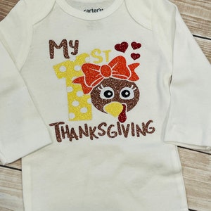 May include: White long-sleeved onesie with the text "My 1st Thanksgiving" in brown glitter. The number 1 is decorated with yellow and white polka dots and a brown turkey head with a red bow.