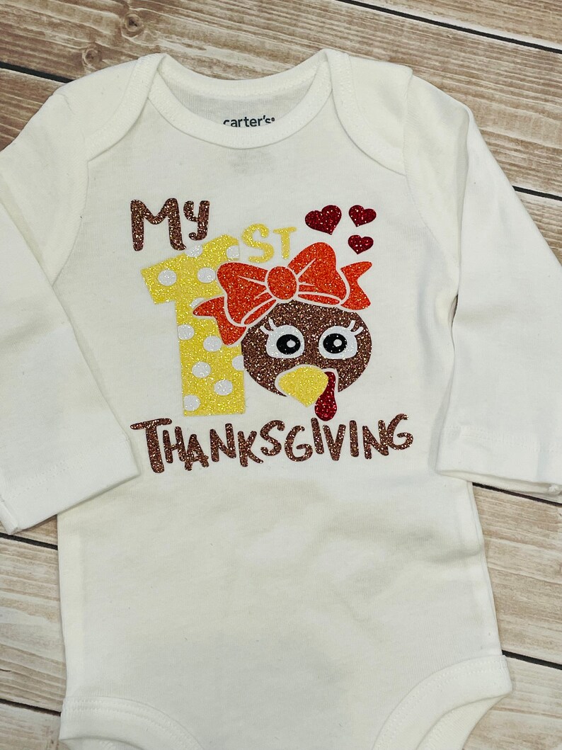 May include: White long-sleeved onesie with the text "My 1st Thanksgiving" in brown glitter. The number 1 is decorated with yellow and white polka dots and a brown turkey head with a red bow.