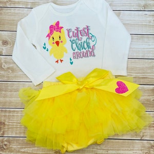 May include: A white long-sleeved onesie with a yellow chick wearing a pink bow and the text "Cutest Chick Around" in pink, blue, and silver glitter. The onesie is paired with a yellow tulle tutu with a pink glitter heart.