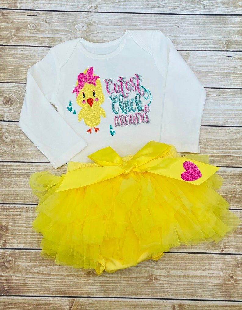 May include: A white long-sleeved onesie with a yellow chick wearing a pink bow and the text "Cutest Chick Around" in pink, blue, and silver glitter. The onesie is paired with a yellow tulle tutu with a pink glitter heart.