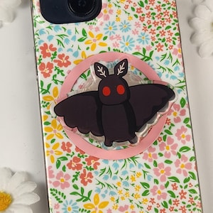 May include: A black moth phone grip with red eyes and a pink circle base is attached to a floral patterned phone case. The case has a white background with pink, yellow, and blue flowers.