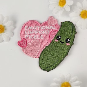 May include: Embroidered patches. One is a pink heart with the text "EMOTIONAL SUPPORT PICKLE", the other is a smiling green pickle with rosy cheeks. The patches are on a white surface with daisy accents.