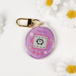 May include: A purple and pink keychain featuring a retro digital pet design. The keychain has a gold-colored clasp and a clear plastic body. The digital pet screen shows a pixelated character. Daisies are in the background.