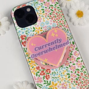 Pode incluir: Uma capa de telefone floral com um suporte de telefone em forma de coração rosa. A pega tem as palavras "Currently Overwhelmed" em roxo. A capa tem um fundo branco com flores vermelhas, rosa, amarelas e azuis.