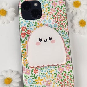 May include: A phone case with a floral design and a white ghost-shaped phone grip. The case has a pattern of pink, red, blue, and yellow flowers. The ghost grip has a cute face with black eyes and a pink blush.
