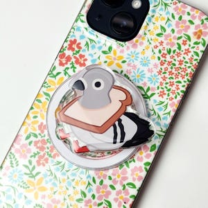 May include: A circular phone grip featuring a cartoon pigeon with a slice of bread around its neck, attached to a smartphone case with a white background and a colorful floral pattern of small pink, yellow, and blue flowers and green leaves.