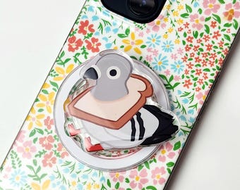 May include: A circular phone grip featuring a cartoon pigeon with a slice of bread around its neck, attached to a smartphone case with a white background and a colorful floral pattern of small pink, yellow, and blue flowers and green leaves.
