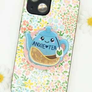 May include: A phone case with a floral pattern and a blue teapot-shaped phone grip. The phone grip has the words "ANXIE TEA" and a tea bag design. The case has a pink border around the grip.