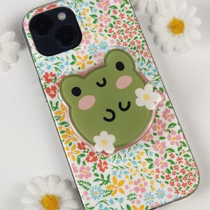 May include: A floral patterned phone case with a green frog-shaped phone grip. The case features a colorful pattern of flowers in pink, blue, and yellow. The frog grip has pink cheeks and white flower accents.