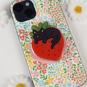 Puede incluir: Una funda de teléfono con estampado floral con un gato negro descansando sobre un popsocket de fresa roja. La funda tiene un fondo blanco con pequeñas flores rosas, azules y amarillas. El popsocket tiene una base rosa.