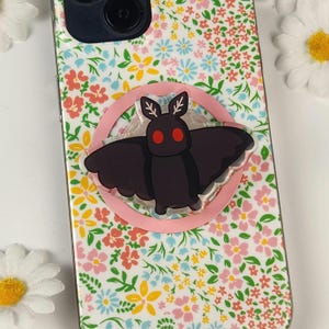 Può includere: Un supporto per telefono a forma di falena nera con occhi rossi e una base circolare rosa è attaccato a una custodia per telefono. La custodia del telefono ha un motivo floreale con fiori rosa, gialli e blu su sfondo bianco.