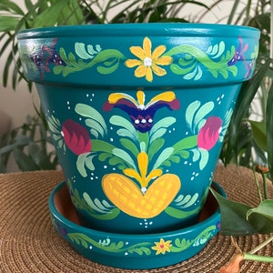 May include: Teal painted terracotta flower pot with a hand-painted design featuring yellow, green, red, and white flowers. The pot has a matching saucer.