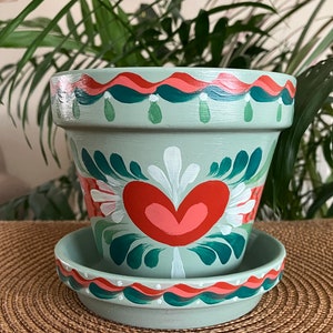 May include: A hand-painted terracotta planter with a green base and a white heart with pink and red accents. The planter has a green and red rim and a matching saucer.