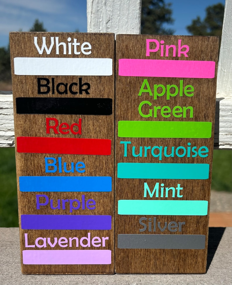 May include: Two wooden signs with color swatches and names. The left sign has the colors white, black, red, blue, purple, and lavender. The right sign has the colors pink, apple green, turquoise, mint, and silver.
