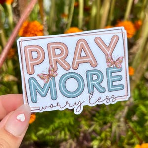 May include: A rectangular sticker with a white background and a thin copper-colored border. The text PRAY MORE is in large, blocky, light orange and teal letters. The words worry less are in a thin, dark brown script font with two butterflies.