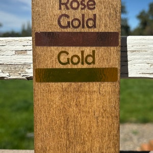 May include: A wooden sample board with three swatches of metallic paint. The top swatch is labeled "Chrome Rose Gold", the middle swatch is labeled "Gold", and the bottom swatch is labeled "Gold".