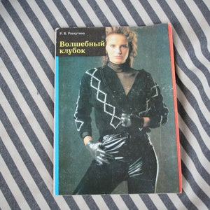 May include: A vintage book cover featuring a woman in a black outfit with a diamond pattern on the sweater. The book title is in Russian, with a blue and red border. The background is a striped fabric.