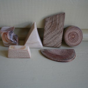 May include: A selection of wooden geometric shapes, including a pyramid, a cylinder, and a semi-circle. The wood pieces have natural grain patterns and vary in colour from light beige to reddish-brown. The shapes are arranged on a light-coloured surface.