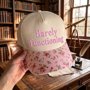 May include: A cream-colored baseball cap featuring the embroidered pink text "Barely Functioning" on the front panel. The brim of the cap is decorated with a pink floral pattern consisting of small red and pink roses with green leaves.