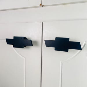 May include: Two black cabinet pulls in the shape of a stylized cross, mounted on a white cabinet door. The pulls have a geometric design with angled edges. The cabinet doors have a simple, classic design.