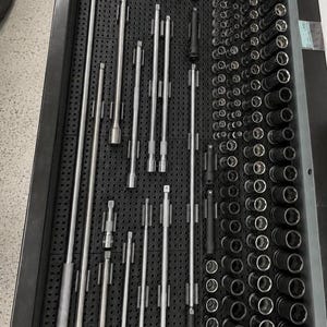 May include: A black tool drawer organizer tray containing a collection of silver metal socket extensions, various small bits, and a large assortment of black and silver metal sockets arranged in neat rows on a perforated pegboard base.