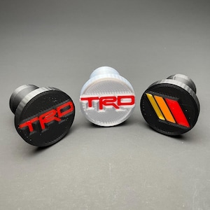 May include: Three circular knob covers with a cylindrical base. The left is black with red TRD text. The center is translucent white with red TRD text. The right is black with three diagonal stripes in yellow, orange, and red.