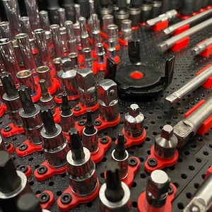 May include: A collection of various metal tools, including sockets, ratchets, and bits, neatly organized on a black surface with red holders. The tools are arranged in rows, showcasing a variety of sizes and shapes, ideal for automotive or mechanical work.