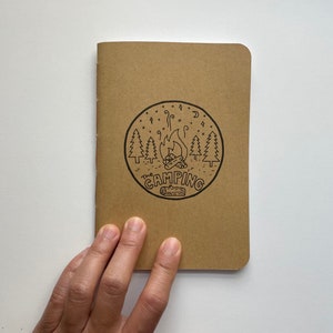 May include: A brown notebook with a black and white illustration of a campfire, trees, and stars. The text "The Camping" is printed in a circle around the illustration.