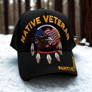 May include: Black baseball cap with gold embroidered text reading "NATIVE VETERAN." The cap features an embroidered design of an eagle, American flag, and feathers. The text "NATIVE" is also embroidered on the brim.