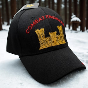 May include: Black baseball cap with "COMBAT ENGINEER" embroidered in red above a gold castle emblem. A small tag with a gold star is attached to the cap. The cap is sitting in the snow.