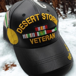 Puede incluir: Gorra de béisbol negra con las palabras "DESERT STORM VETERAN" bordadas en dorado. La gorra presenta cintas militares y una medalla de oro. La visera tiene el texto "ODS 500" en dorado.