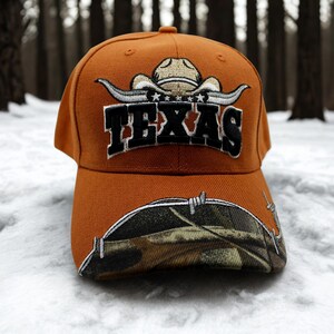 May include: An orange baseball cap with the word "TEXAS" embroidered in black and white. The cap features a cowboy hat and longhorn design above the text. The brim has a camouflage pattern with barbed wire.