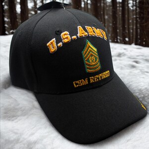 Puede incluir: Gorra de béisbol negra con texto bordado en oro "U.S. ARMY" y un emblema verde y dorado. El texto "CSM RETIRED" está bordado debajo. La gorra se muestra sobre un fondo nevado.