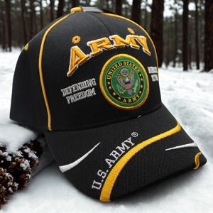 Könnte beinhalten: Schwarze Baseballkappe mit goldenen und weißen Akzenten. Die Kappe zeigt das Wort "ARMY" in Gold, das US Army-Emblem und den Text "U.S. ARMY". Der Schriftzug "Defending Freedom" ist ebenfalls auf die Kappe gestickt.