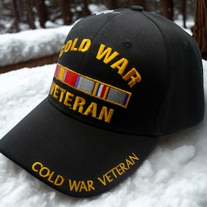 May include: Black baseball cap with the words "COLD WAR VETERAN" embroidered in gold. The cap features a gold embroidered ribbon design with red, white, and blue accents. The cap is sitting on a snowy surface.