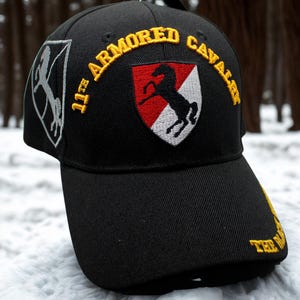 Puede incluir: Gorra de béisbol negra con texto bordado y parches. La gorra presenta el texto "11th ARMORED CAVALRY" en dorado, junto con un escudo y una silueta de caballo. Un parche de caballo está en el lateral. El fondo es nevado.