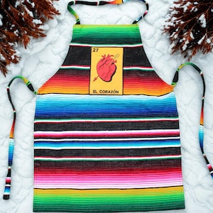 May include: A vibrant apron with a striped pattern in green, blue, red, and yellow. It features a central graphic of a heart pierced by an arrow, with the text "EL CORAZÓN" below. The apron has long ties and neck straps.