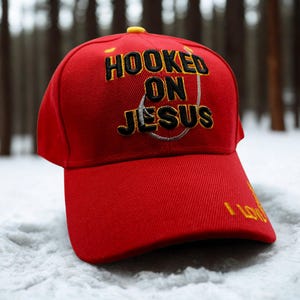 May include: A bright red baseball cap featuring the embroidered black text HOOKED ON JESUS with a silver fishing hook graphic. The cap has a yellow button on the top and the yellow text I LOVE JESUS embroidered on the right side of the curved brim.