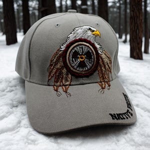May include: Gray baseball cap with an embroidered eagle design. The eagle has white feathers, a yellow beak, and is holding a circular emblem. The words "NATIVE PRIDE" are embroidered on the brim.