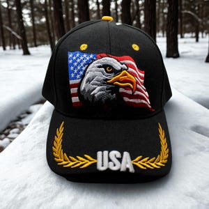 Może przedstawiać: Czarna czapka bejsbolowa z haftowaną amerykańską flagą i wzorem orła bielika. Czapka ma ż&oacute;łty guzik na g&oacute;rze, a słowo "USA" jest wyhaftowane na biało ze złotymi liśćmi laurowymi.