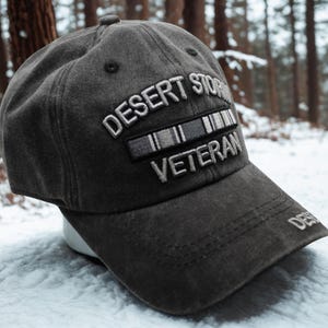 Könnte beinhalten: Eine anthrazitfarbene Baseballkappe mit dem weißen Schriftzug "DESERT STORM VETERAN". Die Kappe hat einen gebogenen Schirm und einen kleinen Knopf oben. Der Hintergrund zeigt einen verschneiten Wald.