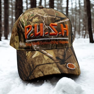 May include: A camouflage baseball cap with the word "P.U.S.H." embroidered in orange, above the text "Pray Until Something Happens." The cap features a brown, green, and tan pattern, and a small orange fish emblem.