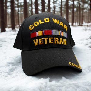May include: A black baseball cap featuring the embroidered yellow text COLD WAR VETERAN. Between the two lines of text is a horizontal embroidered ribbon bar with red, yellow, blue, purple, grey, and white stripes. The word COLD is also on the brim.