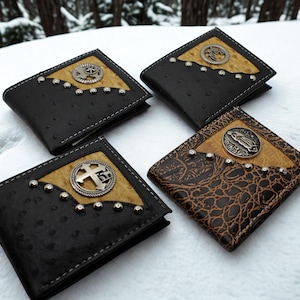 May include: Four bi-fold wallets featuring textured black and brown leather with ostrich-like patterns. Each wallet has a triangular tan inlay accented with silver-toned metal studs and a circular concho emblem depicting a cross or religious figure.