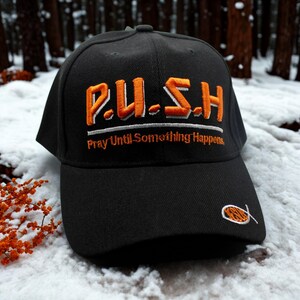 May include: Black baseball cap with the embroidered word "P.U.S.H." in orange, above the phrase "Pray Until Something Happens" in white and orange. A small orange fish symbol is on the side of the cap.
