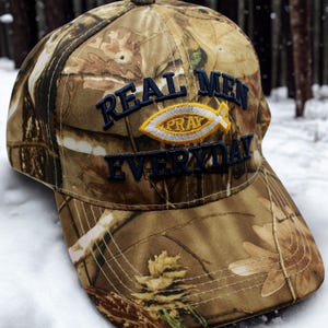May include: Camouflage baseball cap with the text "REAL MEN PRAY EVERYDAY" in navy blue. The cap features a yellow and white fish symbol. The hat is made of a brown and green camouflage pattern.